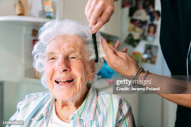 weitwinkelaufnahme einer fröhlichen, niedlichen älteren kaukasischen frau zu hause, die sich von einem professionellen stylisten für häusliche pflegekräfte die haare kämmen lässt - brushing hair stock-fotos und bilder