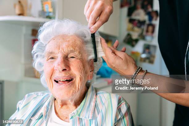 wide angle shot of a cheerful, cute elderly senior caucasian woman at home having her hair combed by a professional in-home caregiver stylist - haardracht stockfoto's en -beelden