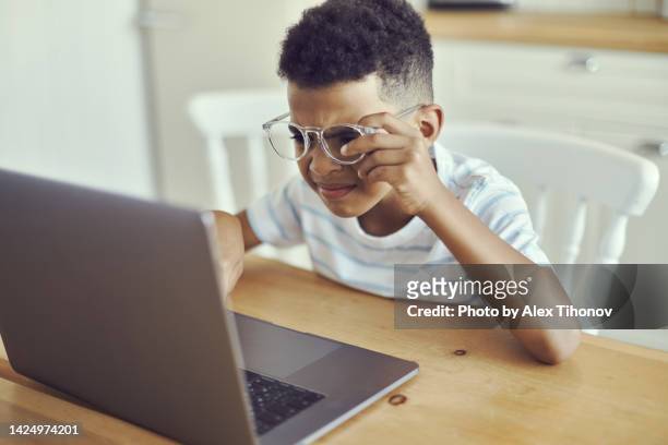 little african kid boy in spectacles looks at the laptop screen, squints, try to read information, does not see, has vision problems. visual impairment due to excessive usage of modern wireless tech concept - entrecerrar los ojos fotografías e imágenes de stock