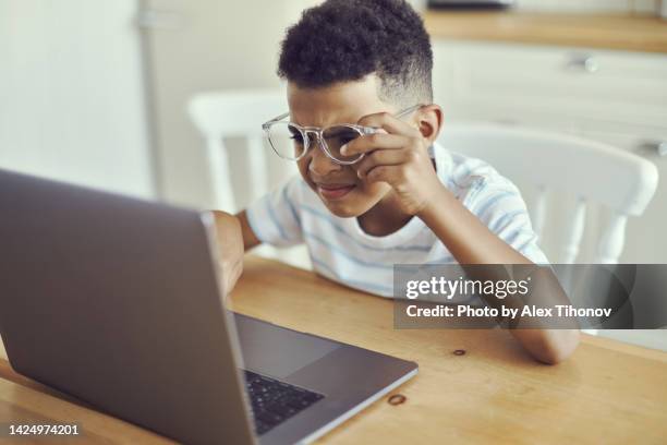 little african kid boy in spectacles looks at the laptop screen, squints, try to read information, does not see, has vision problems. visual impairment due to excessive usage of modern wireless tech concept - miopia foto e immagini stock