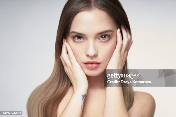 beauty portrait of a beautiful young woman on a light grey background with flowing long hair, good skin and tattoos on her arms. shirtless blonde woman holding hands near her face, nude makeup and natural beauty, skin care, hair care - couleur chair photos et images de collection