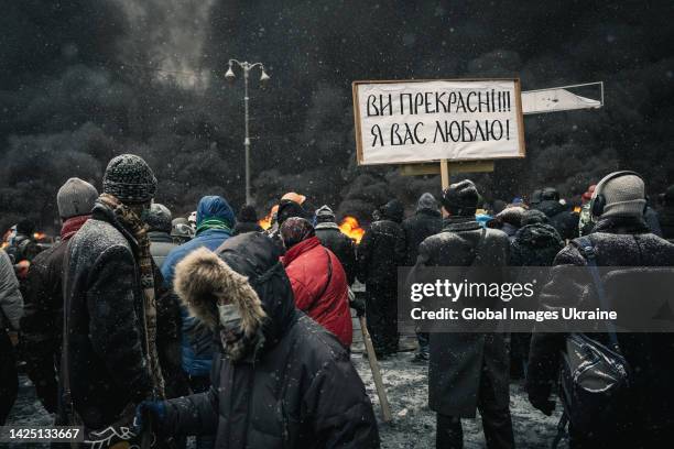 Oleksandr Melnyk, Ukrainian muralist, holds a banner “You’re beautiful!!! I love you!” standing amid protesters in front of rising smoke from burning...