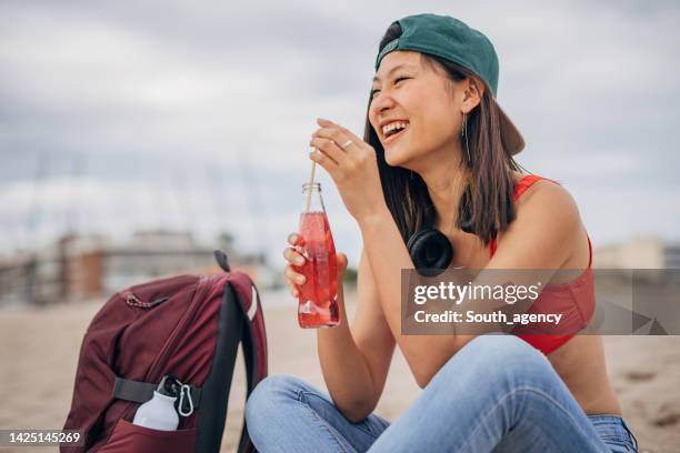 junge chinesin, die an einem strand sitzt und soda trinkt - sodawasserflasche stock-fotos und bilder