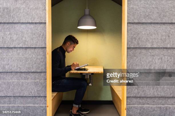 young man sitting in small cubicle typing on digital tablet - office cabin stockfoto's en -beelden