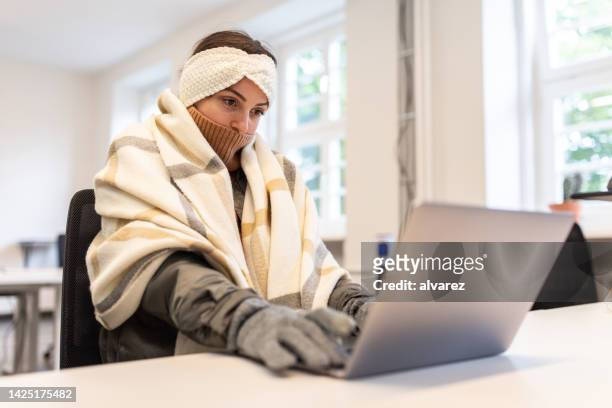 mujer sintiéndose fría cubierta con una manta usando una computadora portátil en la oficina - frio fotografías e imágenes de stock