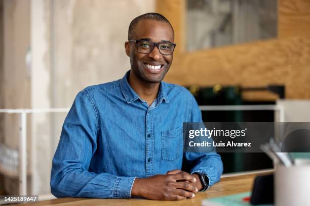 portrait of a smiling young african businessman sitting at desk - black shirt stock pictures, royalty-free photos & images