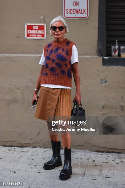 Grece Ganhem is seen wearing brown sunglasses, white t-shirt, a dark brown with navy blue print pattern sleeveless wool sweater, a brown pleated midi...