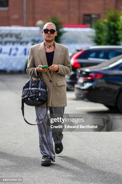 Guest wears black sunglasses, a black with brown pendant necklace, a gold pendants necklace, a black t-shirt, a brown and black checkered print...