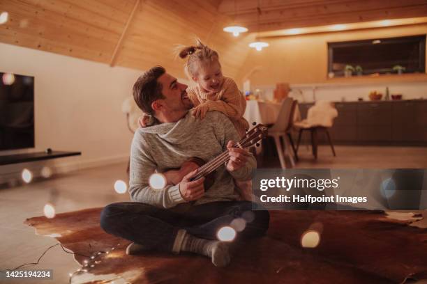 young cheerful dad sitting with his daughter in living room and playing on guitar. - cosy family stock pictures, royalty-free photos & images