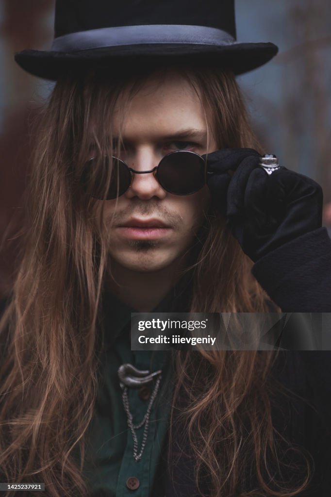 Gothic man with long hair in glasses and hat. Black classic clothes. Vampire in black suit on autumn background. Halloween costume. Portrait