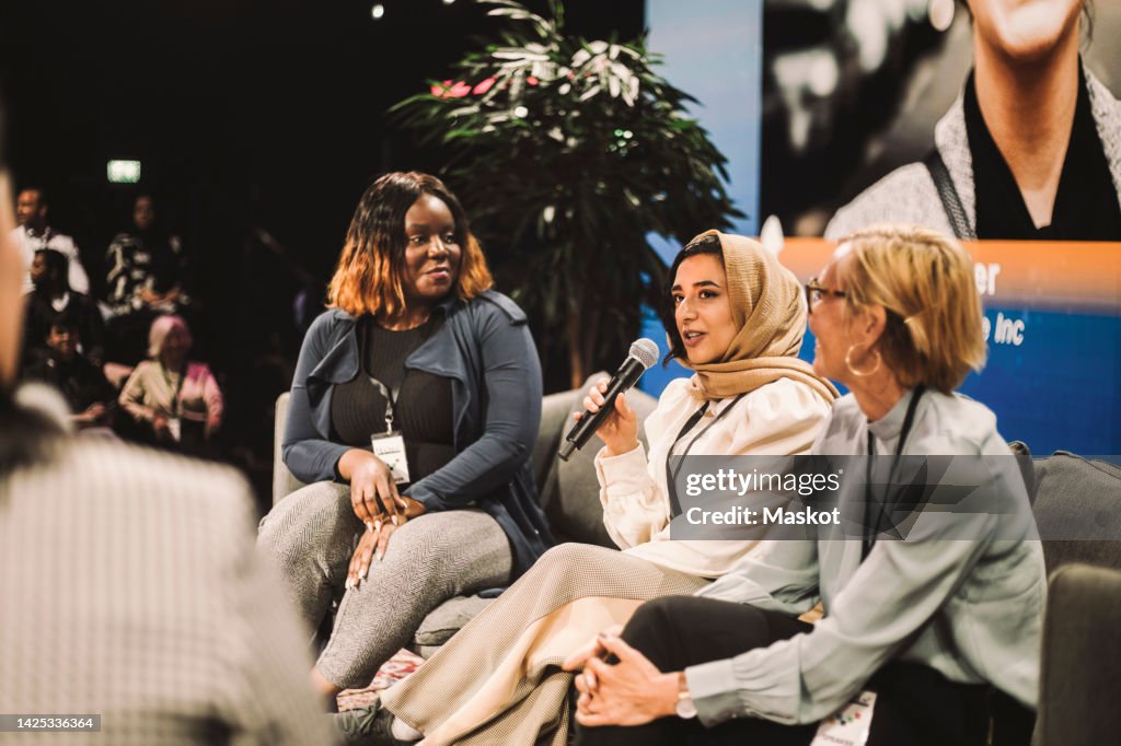 Smiling female entrepreneur looking at colleague interacting with interviewer during panel in convention center