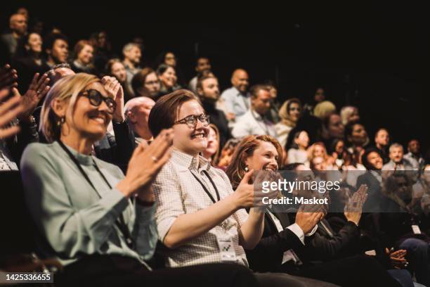 happy colleagues applauding while sitting in conference event at convention center - audience - fotografias e filmes do acervo