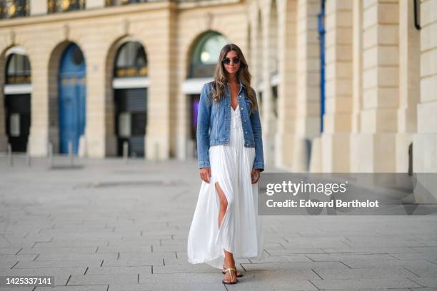 Amanda Derhy wears earrings, sunglasses, a blue denim jacket, a white summer maxi pleated low-neck slit off-shoulder dress, sandals, during a street...