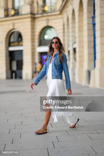 Amanda Derhy wears earrings, sunglasses, a blue denim jacket, a white summer maxi pleated low-neck slit off-shoulder dress, sandals, during a street...