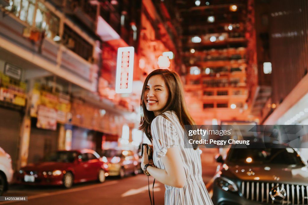 Young cheerful Asian woman looking back in front of the neon lights of Hong Kong on the streets at night while holding her camera