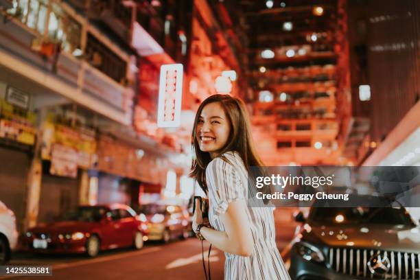 young cheerful asian woman looking back in front of the neon lights of hong kong on the streets at night while holding her camera - chinese people stock pictures, royalty-free photos & images