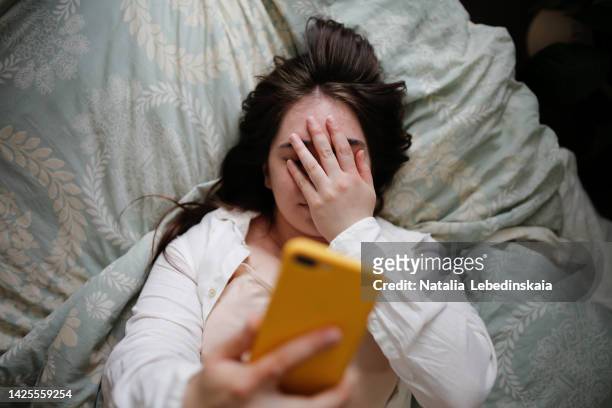 sleepy overweight young woman with flowing hair using phone on bed, close face palm. top view - embarrassment foto e immagini stock