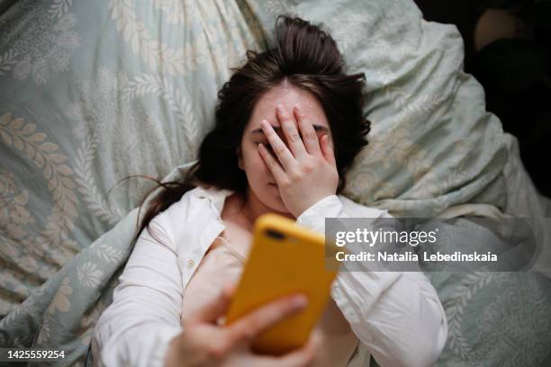 sleepy overweight young woman with flowing hair using phone on bed, close face palm. top view - polshorloge stockfoto's en -beelden