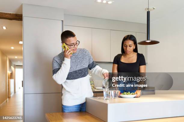 a bespectacled man talking on the phone and pouring two glasses of water and next to him a short-haired woman holding a salad - plain salad stock pictures, royalty-free photos & images