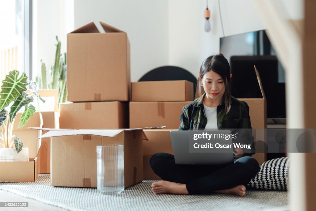 Young woman using laptop while sitting on the floor in new home with cardboard boxes