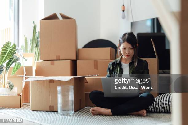 young woman using laptop while sitting on the floor in new home with cardboard boxes - rent assistance stock pictures, royalty-free photos & images