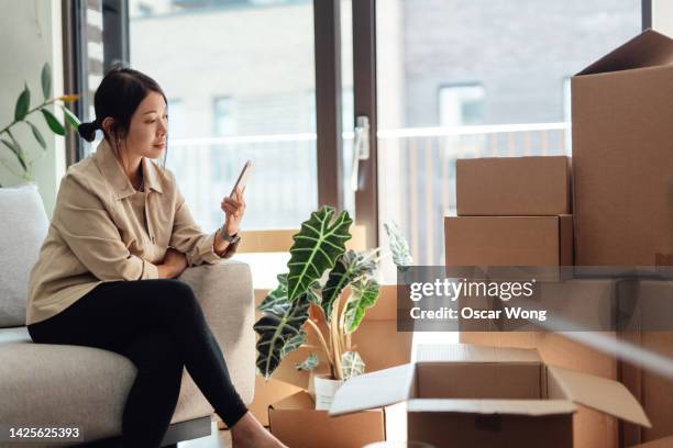 young woman using smart phone while sitting on sofa in new home with cardboard boxes - rental assistance stock pictures, royalty-free photos & images