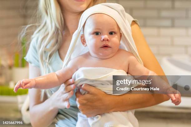 young adult mother is giving her infant son a bath in the kitchen sink at home and wrapping him in a towel - baby bath toys stock pictures, royalty-free photos & images