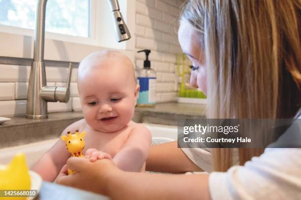 young adult mother is giving her infant son a bath in the kitchen sink at home and wrapping him in a towel - baby bath toys stock pictures, royalty-free photos & images