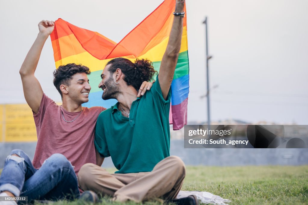 Männliches schwules Paar, das auf Gras sitzt und die Schwulenflagge hält