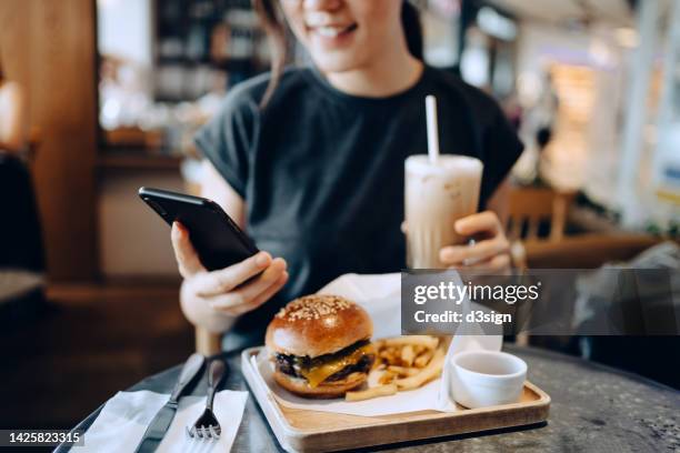 cropped shot of smiling young asian woman using smartphone while enjoying her lunch, cheeseburger with french fries and iced coffee in a stylish cafe. people, food, lifestyle and technology concept - fastfoodrestaurant stockfoto's en -beelden