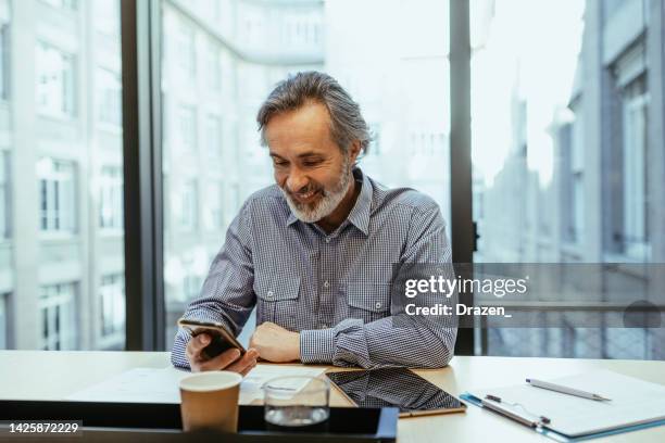 grauhaariger mitarbeiter, der im büro arbeitet und auf das smartphone schaut, während er die mobile app verwendet - old wise business man white stock-fotos und bilder