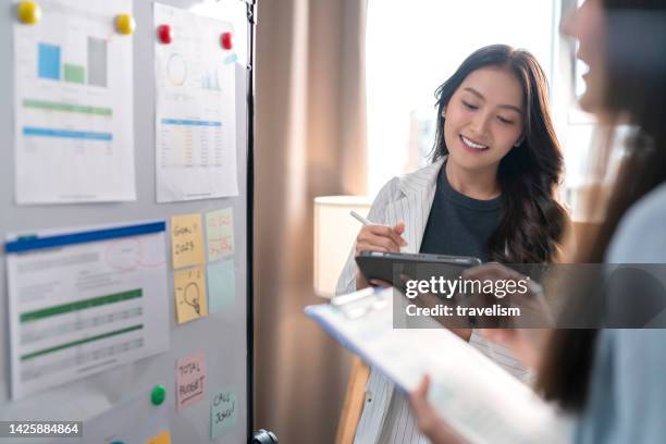 zwei geschäftsfrauen, die mit ihrer kollegin ideen auf whiteboard brainstormen. weibliche geschäftspartner, die eine brainstorming-sitzung über das finanzbudget im startup-besprechungsraum haben. - team looking at adhesive notes in board room during meeting stock-fotos und bilder