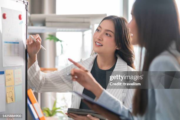 two businesswoman brainstorming ideas on whiteboard with colleague. female business partners having brainstorming session about financial budget in startup meeting room. - showing pen stock pictures, royalty-free photos & images