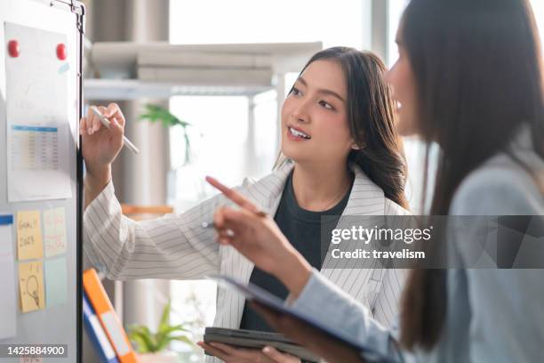 two businesswoman brainstorming ideas on whiteboard with colleague. female business partners having brainstorming session about financial budget in startup meeting room. - whiteboard session stock pictures, royalty-free photos & images