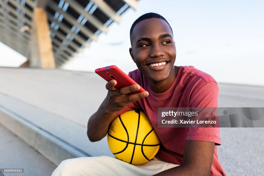 Happy young black man with basquetball ball relaxing outdoors while using smart mobile phone to send an audio voice message - Technology, youth and millennial generation people concept