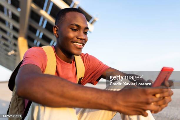 cheerful young adult black hispanic latin man having fun using smart mobile phone sitting outdoors at sunset - technology, social media and millennial people concept - boys relaxing stock-fotos und bilder