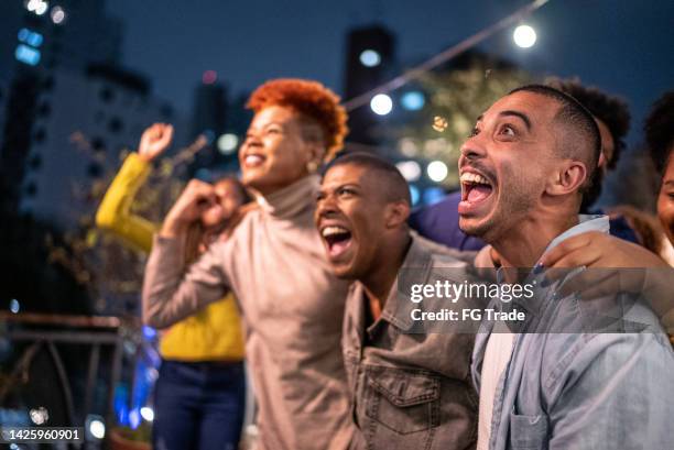 sports fans watching a match and celebrating at a bar rooftop - partida desporto imagens e fotografias de stock