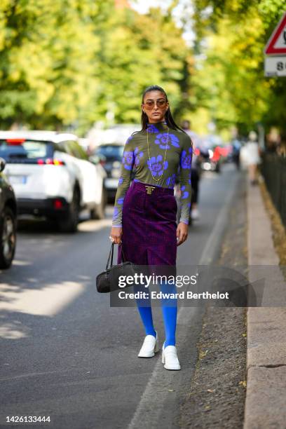 Melanie Darmon wears brown sunglasses, gold earrings, a gold long chain necklace, a khaki and purple print pattern high neck / long sleeves body,...