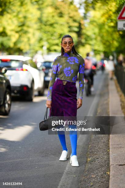 Melanie Darmon wears brown sunglasses, gold earrings, a gold long chain necklace, a khaki and purple print pattern high neck / long sleeves body,...