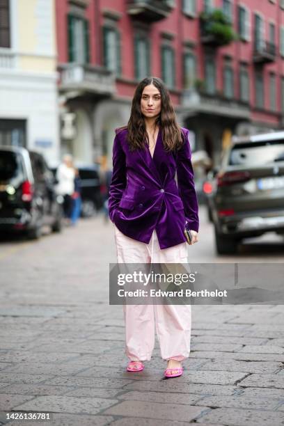 Erika Boldrin wears a dark purple velvet oversized jacket, pale pink puffy baggy pants, neon pink shiny leather strappy heels sandals , outside...
