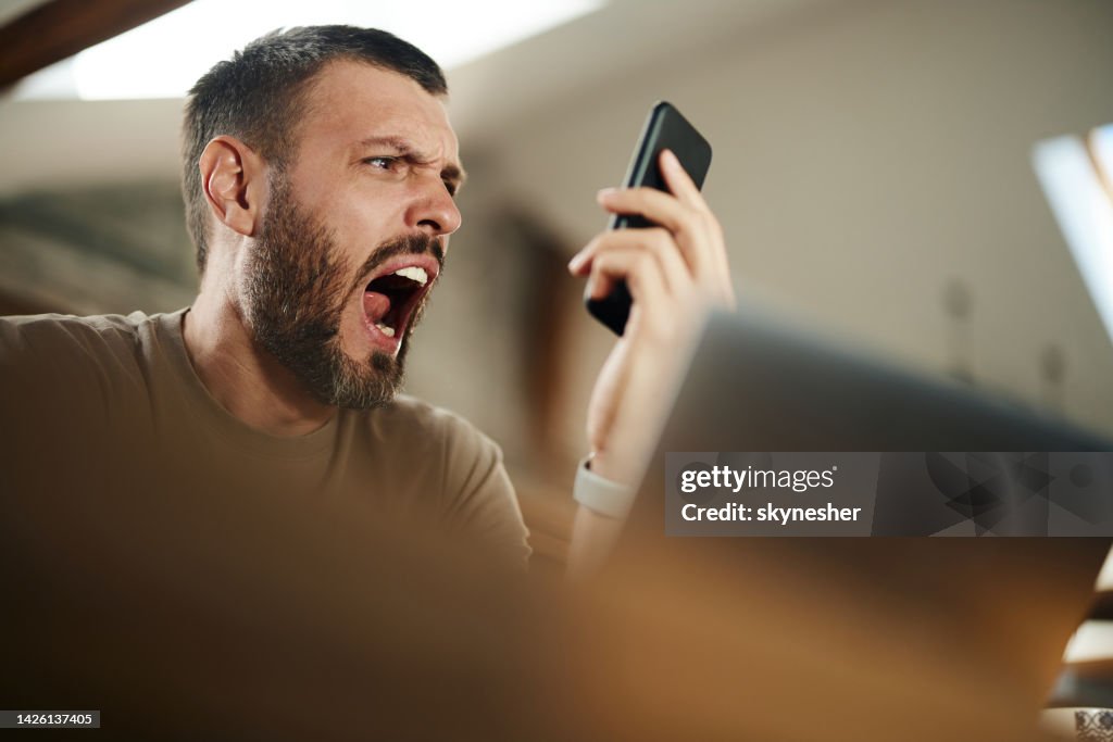 Homme en colère qui crie en utilisant un téléphone portable tout en travaillant à la maison.