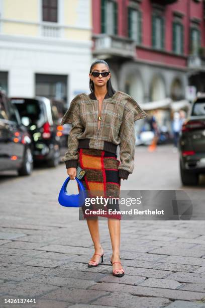 Melanie Darmon wears a gold hair clip, black sunglasses, gold earrings, a brown with blue and red checkered print pattern oversized jacket, a royal...