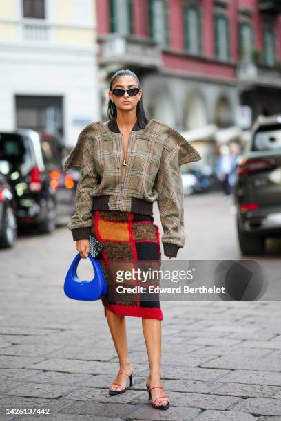 Melanie Darmon wears a gold hair clip, black sunglasses, gold earrings, a brown with blue and red checkered print pattern oversized jacket, a royal...