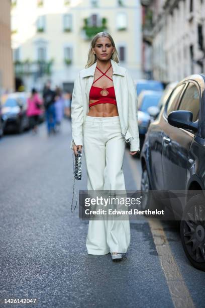 Caroline Daur wears a red strappy neck / cut-out / cropped top, a white latte jacket, matching white latte shiny leather flared pants, a silver...