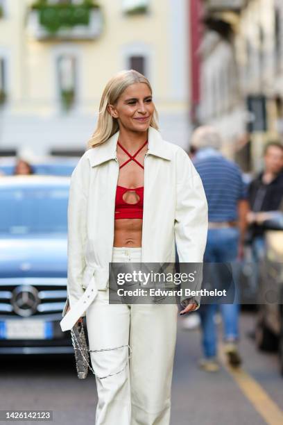 Caroline Daur wears a red strappy neck / cut-out / cropped top, a white latte jacket, matching white latte shiny leather flared pants, a silver...