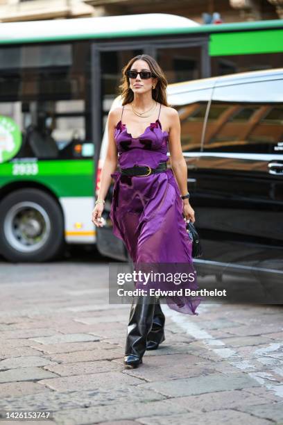 Tamara Kalinic wears black sunglasses, gold earrings, gold chain necklaces, a purple ruffled silk V-neck / tank-top midi dress, a black shiny leather...