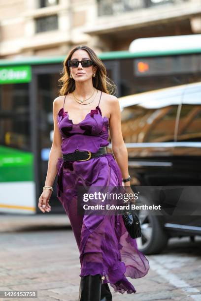 Tamara Kalinic wears black sunglasses, gold earrings, gold chain necklaces, a purple ruffled silk V-neck / tank-top midi dress, a black shiny leather...