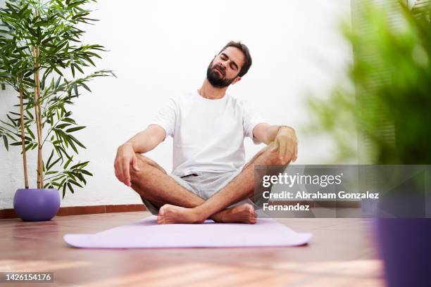 young man sitting on a mat practicing yoga stretching his neck - rückentraining stock-fotos und bilder