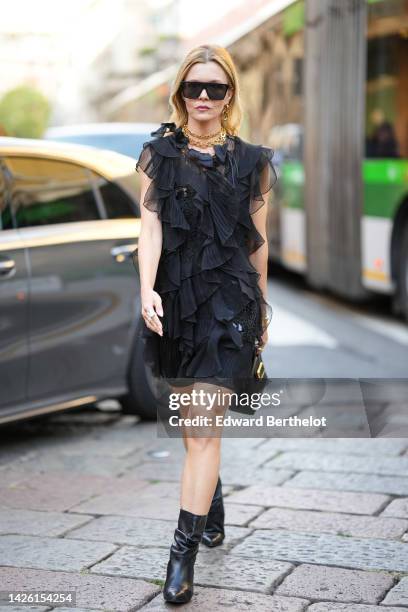 Elizabeth Sulcer wears black sunglasses, gold earrings, gold large chain necklaces , a black ruffled tulle/ embroidered pearls / sleeveless / short...