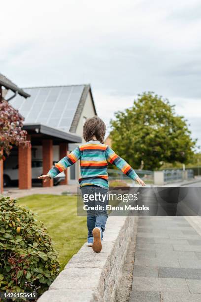 view from the back of a little boy in a striped sweater, who is walking along a small fence next to the house with his arms spread out to the sides - sides 1 2 stock pictures, royalty-free photos & images